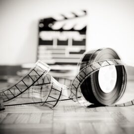Image en noir et blanc montrant une bobine de film au premier plan et clap de cinéma au second plan posés sur un parquet en bois.
