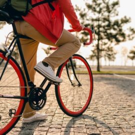gros plan sur les jambes en baskets et les mains sur le volant d'un homme en sweat rouge et pantalon beige sur un vélo avec un sac à dos