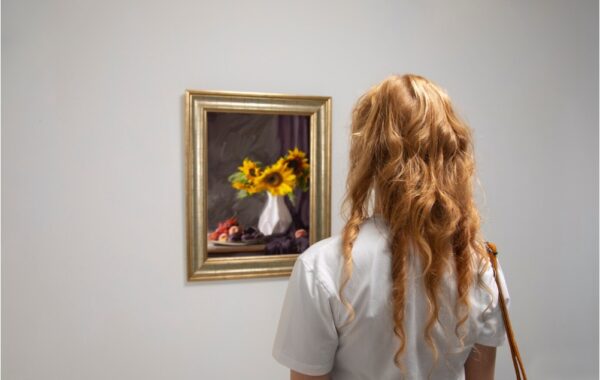 Femme aux cheveux bouclés et avec un tee-shirt blanc observant un tableau qui représente des tournesols dans un vase.