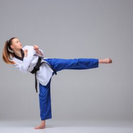 Jeune fille habillée en tenue d'arts martiaux et faisant un coup de pied devant elle. Elle porte un haut de kimono blanc, un bas bleu et une ceinture noire