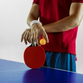 Homme en tenue de sport qui pratique le ping-pong. Il a les mains croisées sur sa raquette posée sur la table de ping pong.